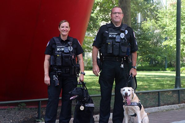 K9 units posing in front of large red cylindrical sculpture, officer Wesley on left with canine Uman, a black labrador retriever, and Sgt. Mackey on the right with canine Zzisa, a yellow labrador retriever.