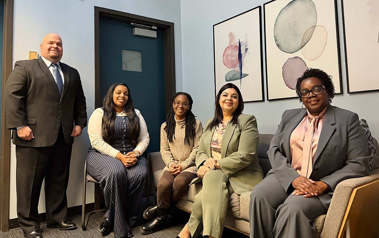 The Special Services team sits in a waiting area on a sofa, chair, and standing. They are all facing the camera and smiling. The background is blue with soft pastel artwork on the wall behind them.